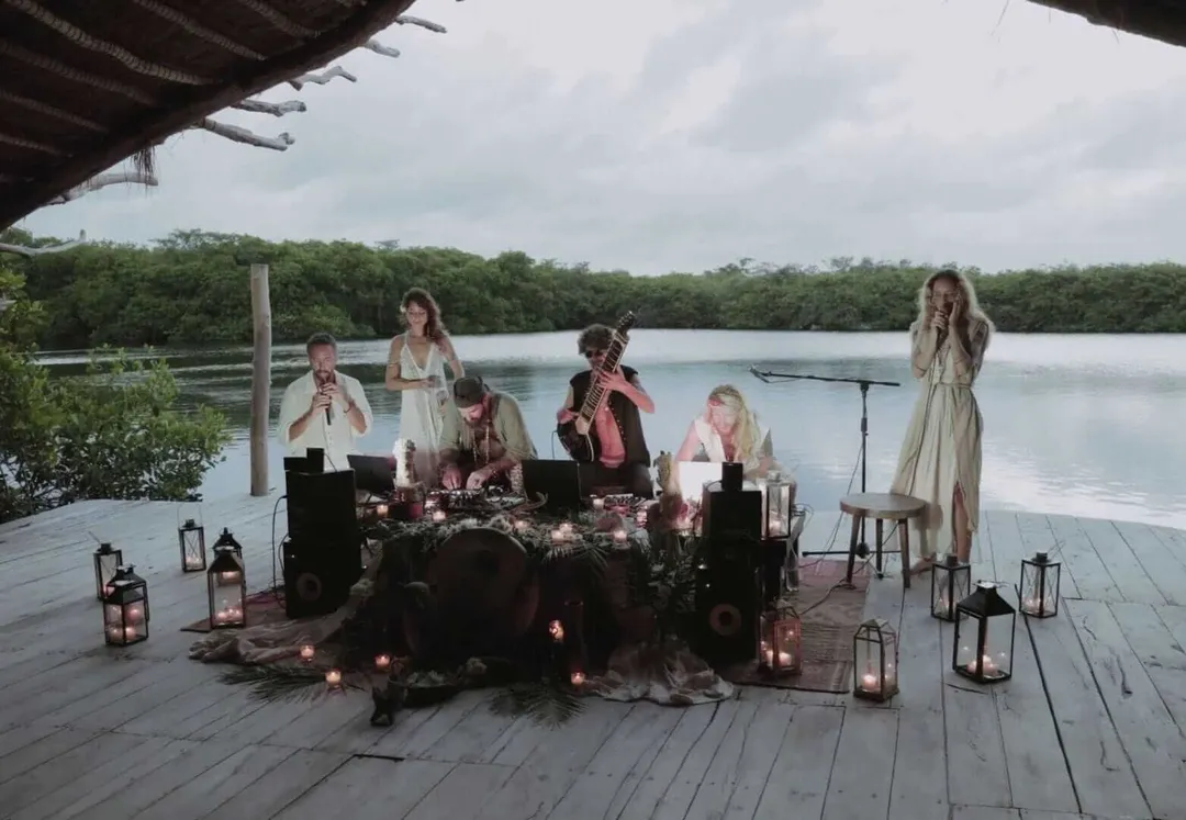 A musical group performs on a wooden dock overlooking a calm waterway surrounded by mangrove trees, with candles and lanterns arranged on the stage
