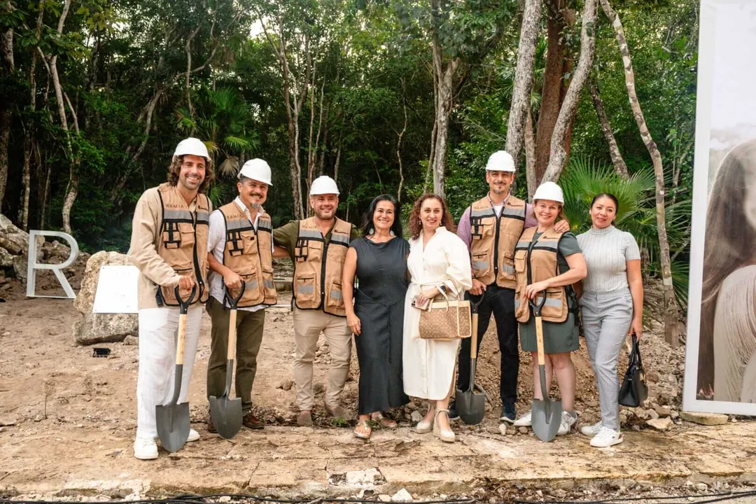 Group of people wearing hard hats and work vests stand together on a construction site surrounded by tropical trees