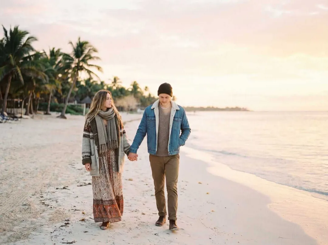 Two people walk holding hands along a beach at sunset, wearing jackets and warm layers despite tropical palm trees lining the shore