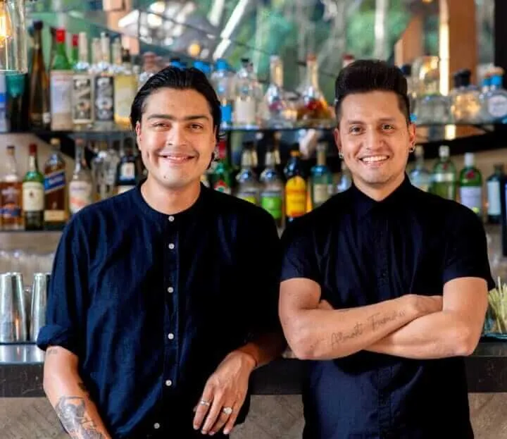 Two bartenders in dark shirts stand behind a bar counter with bottles of liquor displayed on shelves behind them