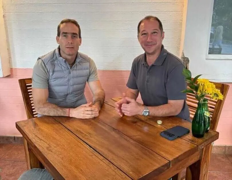 Two men sit at a wooden table during an indoor interview, with a vase of yellow flowers and a phone visible on the table behind them