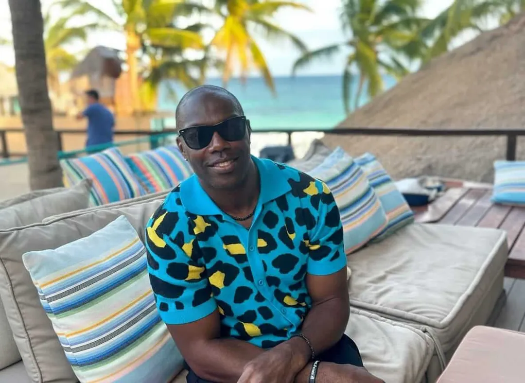 La Zebra Tulum Hosts Sold-Out Super Bowl Celebration With Terrell Owens Man in blue patterned shirt and sunglasses sits on lounge chairs at a beachfront venue with palm trees and ocean visible in the background