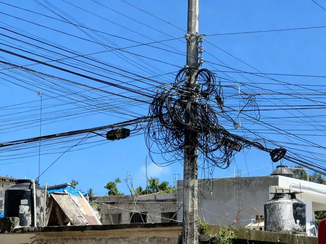 Tangled electrical wires and cables cluster around a utility pole above residential buildings under clear sky