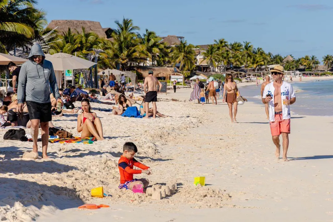 Beachgoers relax and play in the sand and surf at a crowded beach resort with thatched-roof structures and palm trees lining the shore