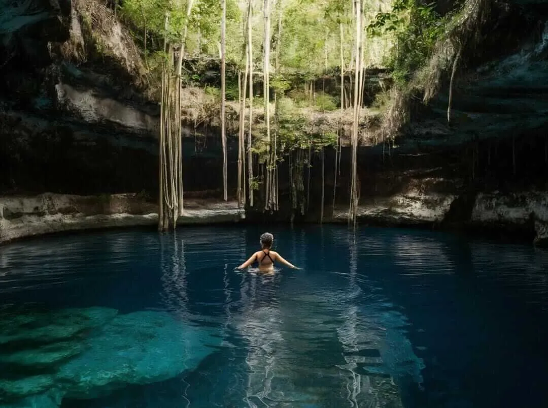 A swimmer floats in clear blue water inside a cenote surrounded by limestone cliffs and hanging vines