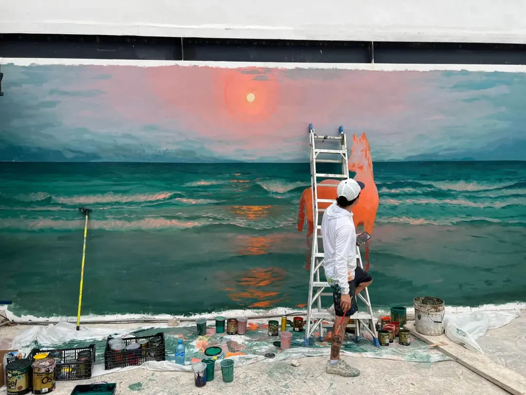 Artist on a ladder paints a large mural depicting an ocean sunset scene on a wall, with paint supplies and tools scattered on the ground below
