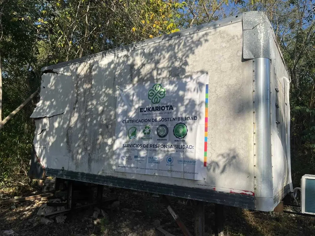 A white cargo trailer with sustainability certification signage and logos sits among trees and brush in an outdoor setting