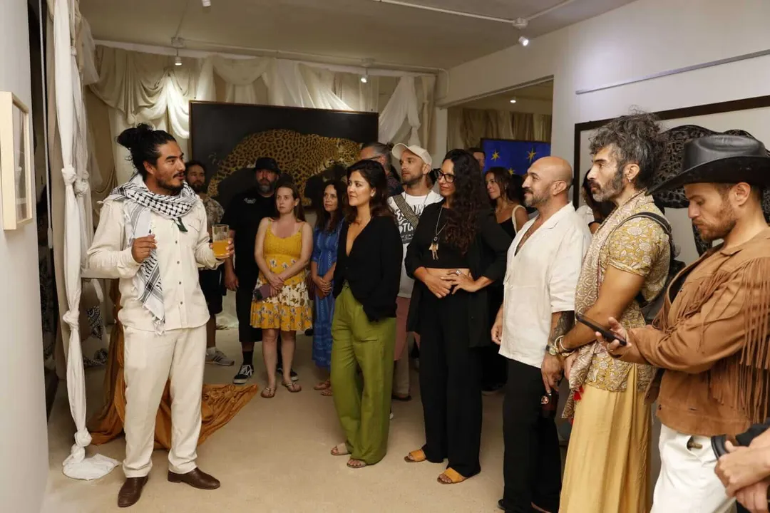 Encierros by Luis Antonio Trujillo draws record crowd in Tulum A man in a cream suit and patterned scarf speaks to a gathered crowd inside an art gallery space with draped curtains and artwork on the walls