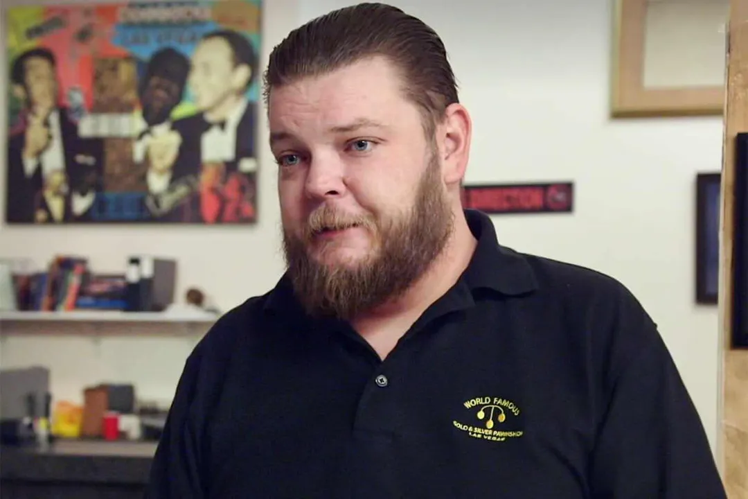 Man with beard wearing a black polo shirt with embroidered logo speaks indoors with shelving and artwork visible in the background