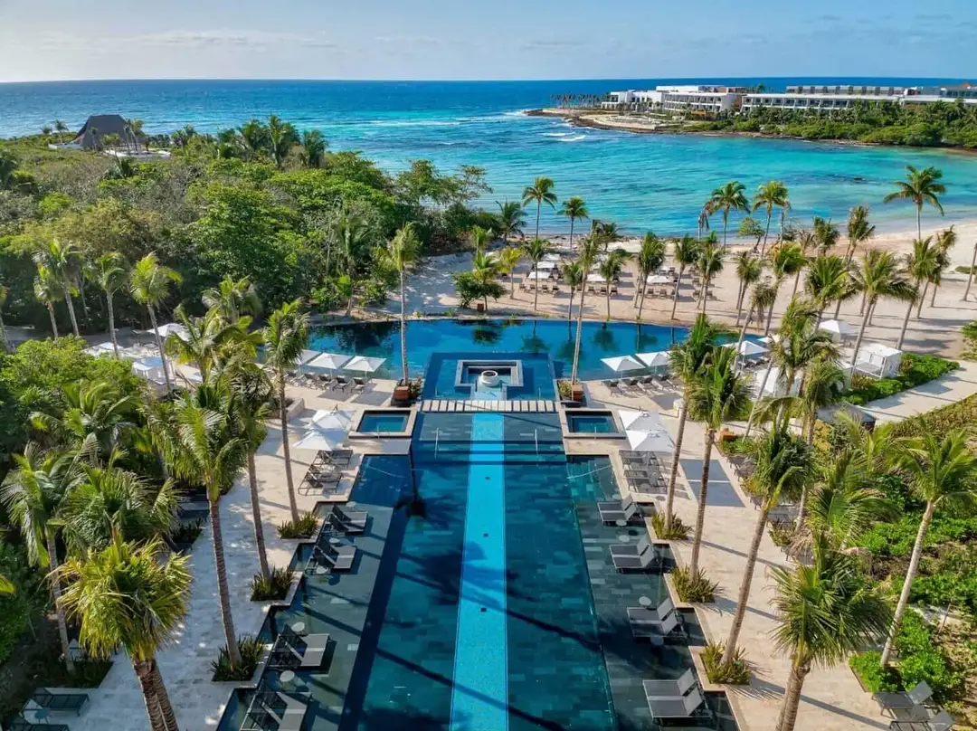 How the Conrad Tulum Invited Us In and Made Us Never Want to Leave Aerial view of a beachfront resort with a long swimming pool surrounded by palm trees, lounge chairs, and a sandy beach with turquoise water beyond