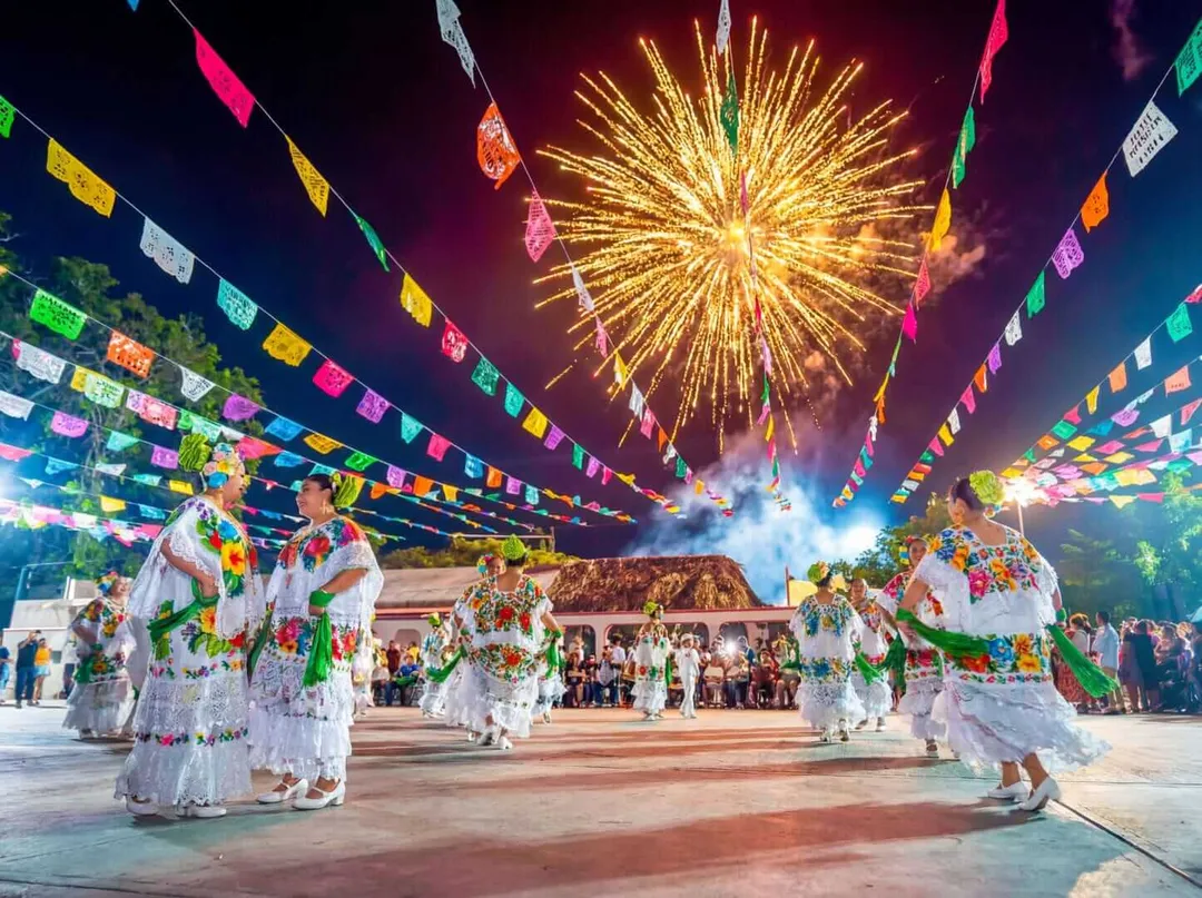 Performers in white embroidered dresses dance in a courtyard decorated with colorful paper banners as fireworks burst overhead near a temple structure