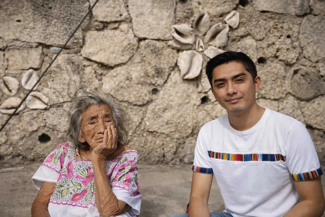 The woman who welcomed generations into Tulum’s families An elderly woman and a younger man sit together against a weathered stone wall with decorative shells