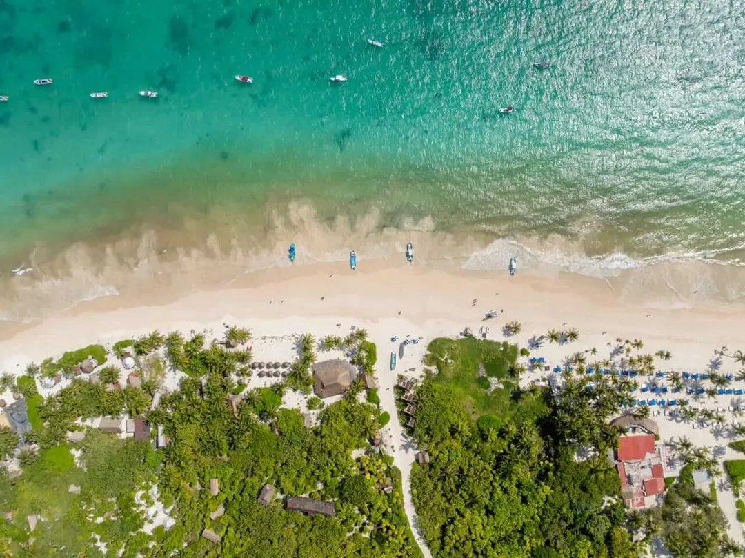 Aerial view of a beach with turquoise water, white sand, waves, and beachgoers, bordered by dense green vegetation and resort structures along the shoreline