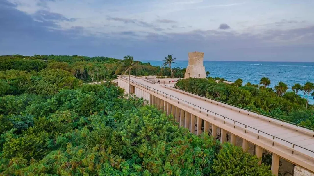 Foreign Residents Embrace Jaguar Park’s Free Access in Tulum A wooden boardwalk with railings extends through dense green vegetation toward a stone tower on a coastal bluff overlooking the sea