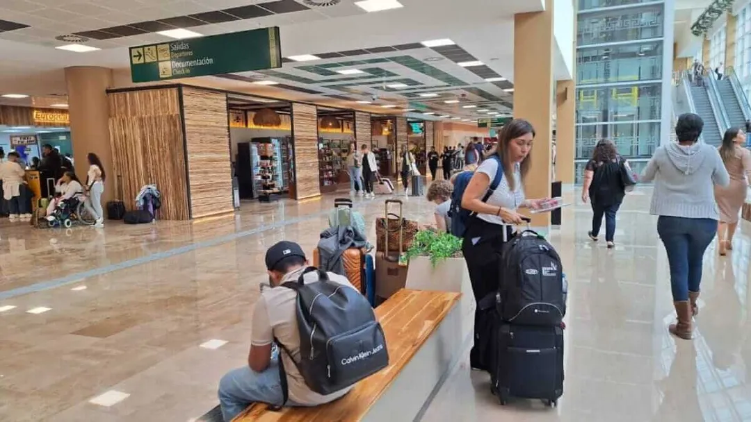 Travelers with luggage sit and stand in a modern airport terminal with retail shops and seating areas