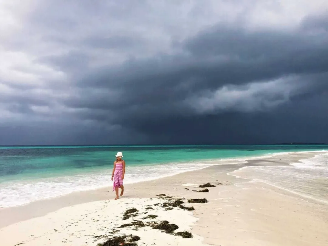 Person in pink clothing and hat standing on sandy beach facing turquoise water under dark storm clouds