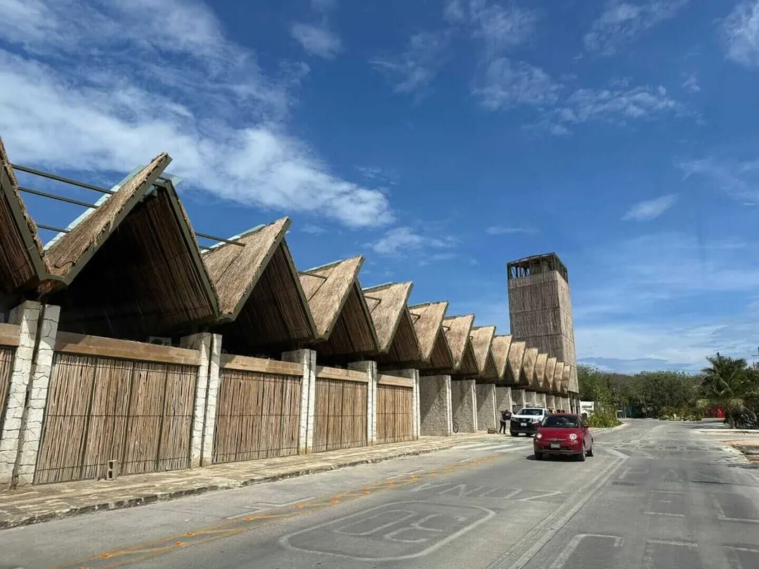 Tulum Commercial Rental Prices Soar, Emptying Jaguar Park Shops A row of wooden-roofed commercial structures with concrete pillars stands empty along a paved road, with a red vehicle parked nearby and a tower visible in the