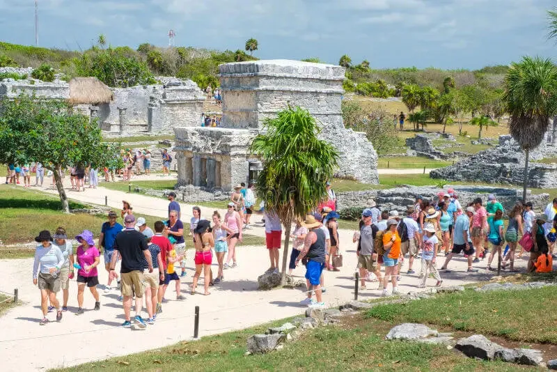 What the Mexico Travel Advisory Doesn’t Tell You Tourists explore ancient stone ruins with palm trees and grassland surrounding the archaeological site