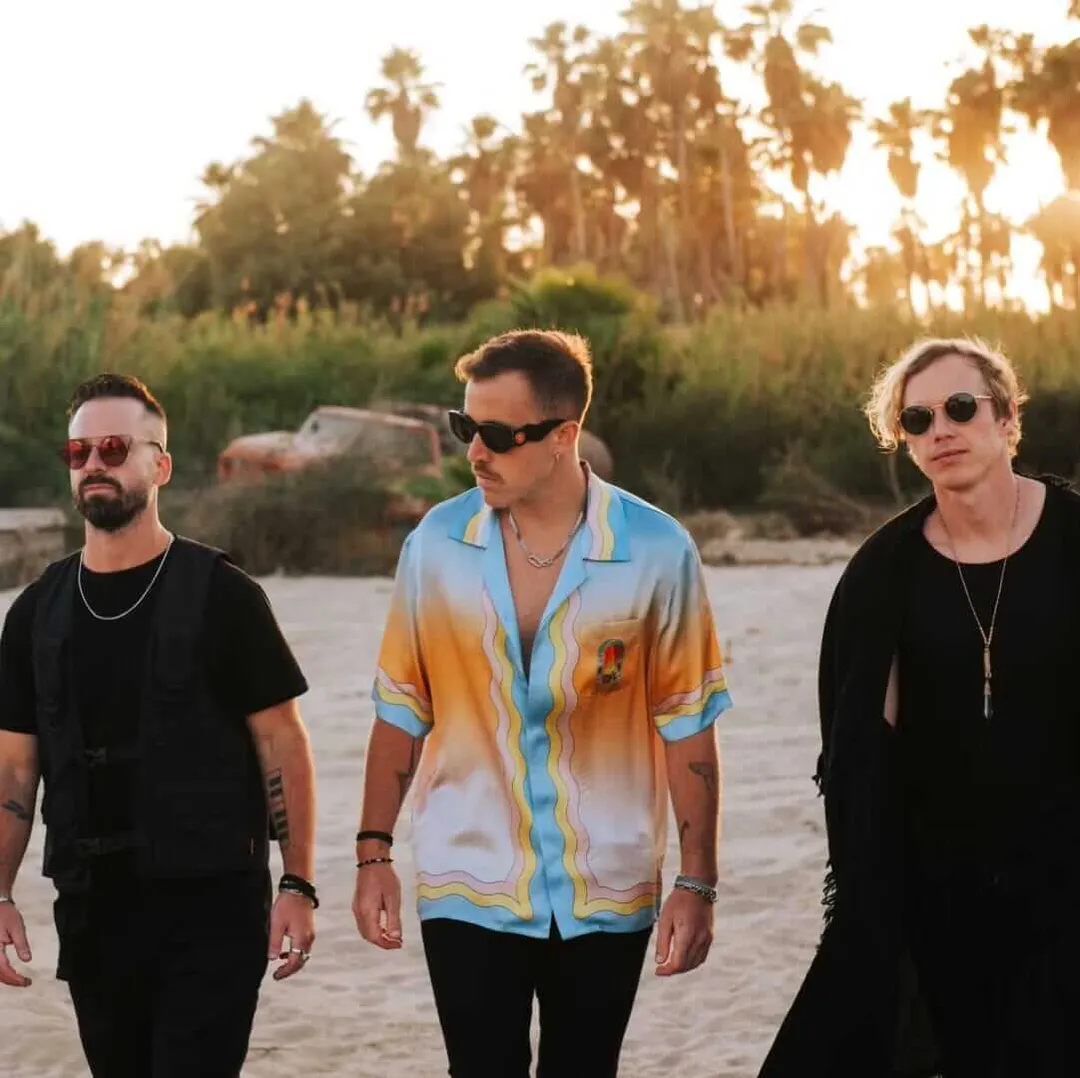 RÜFÜS DU SOL Return to Tulum for Exclusive DJ Set at Zamna 2026 Three men wearing sunglasses walk along a beach at sunset with palm trees and vegetation in the background