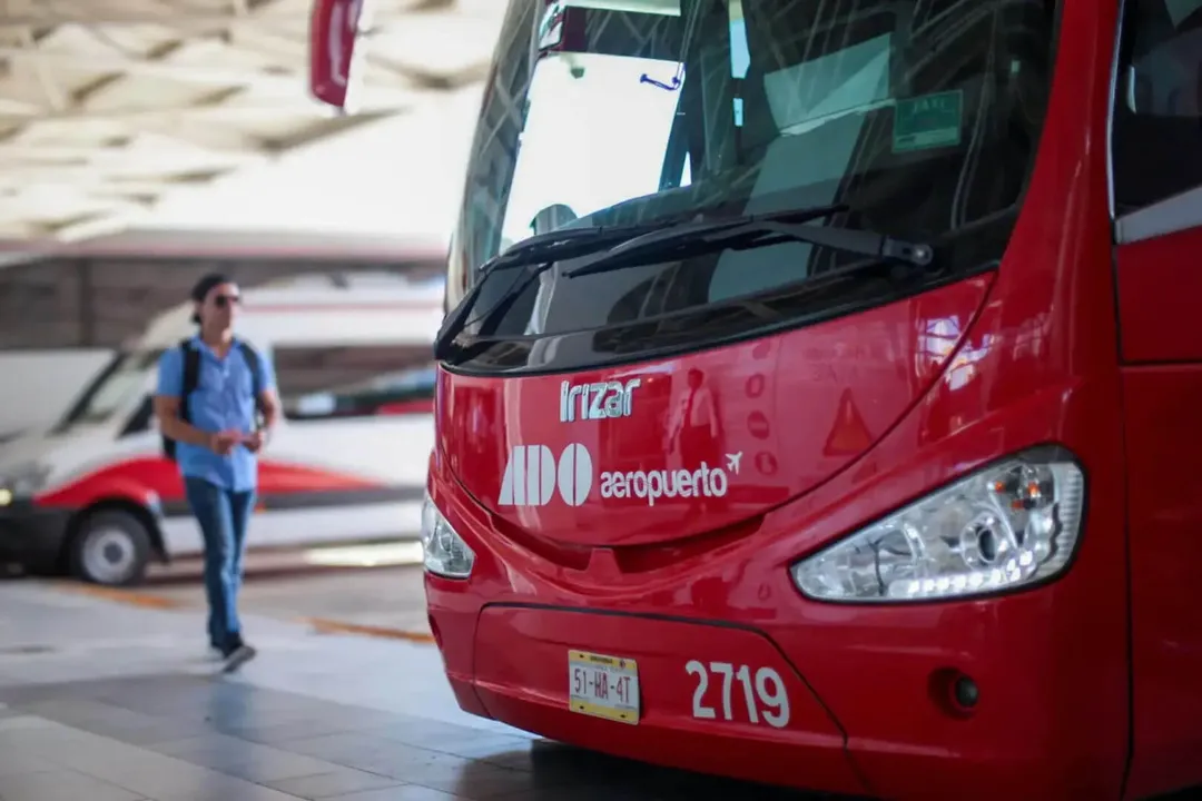 Red shuttle bus parked at an airport terminal with a person in a blue shirt walking nearby