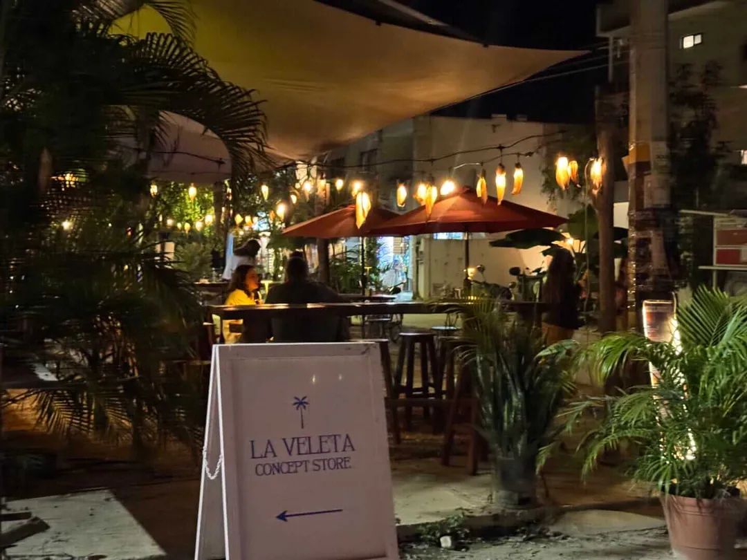 Outdoor dining area at night with string lights, potted plants, and a red umbrella, with a sign reading "La Veleta Concept Store" visible in the foreground