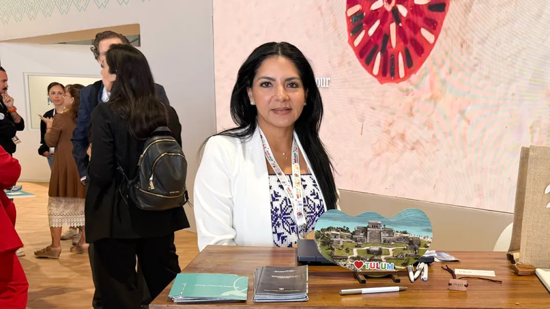 Tulum Reactivates Tourism Councils as Businesses Warn of a Growing Crisis Woman wearing a white blazer and credential badge sits at a table with tourism materials during an indoor event with other attendees in the background