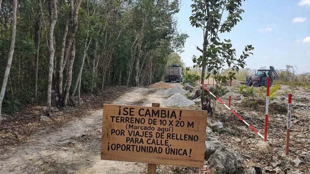 Wooden sign advertising land for sale stands along a dirt road lined with trees, with construction equipment and a truck visible in the background