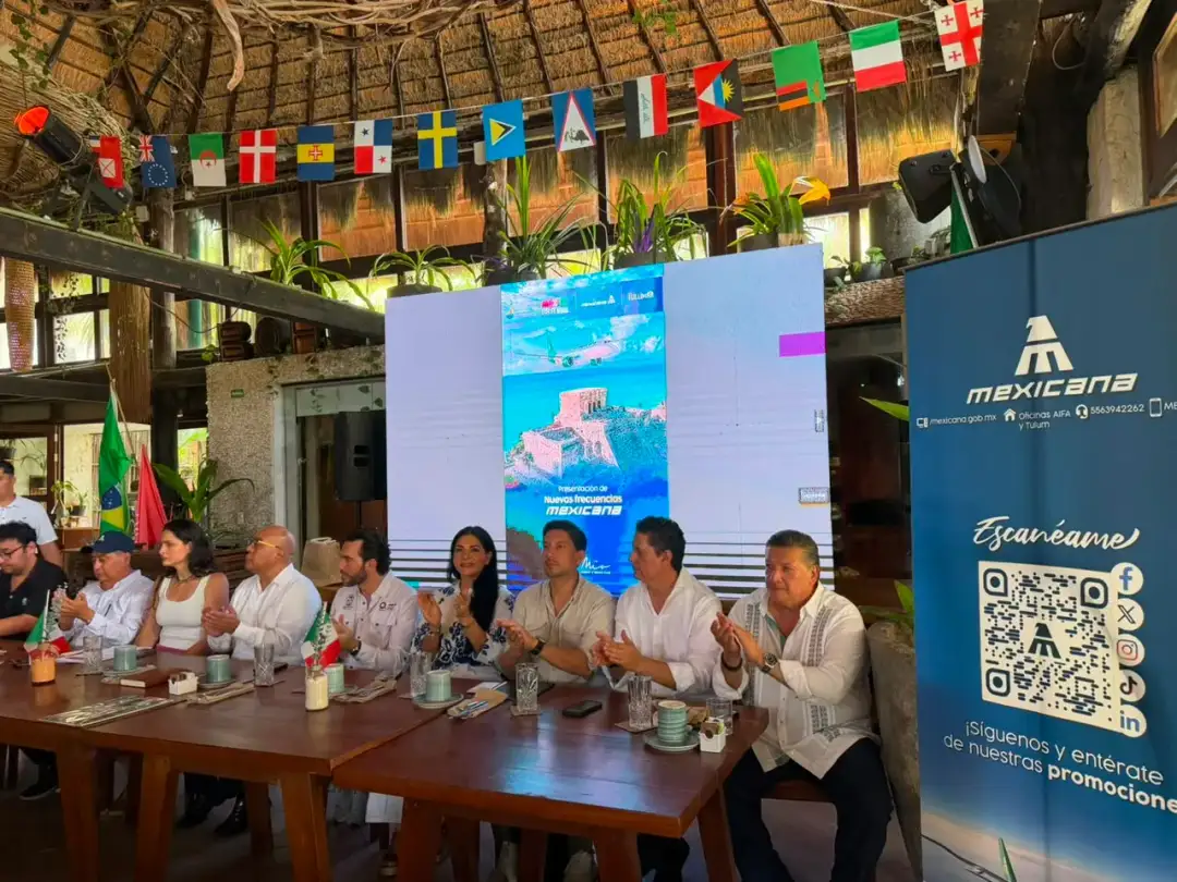 Group of people seated at a long table during an event with international flags hanging overhead and promotional screens displaying tourism imagery