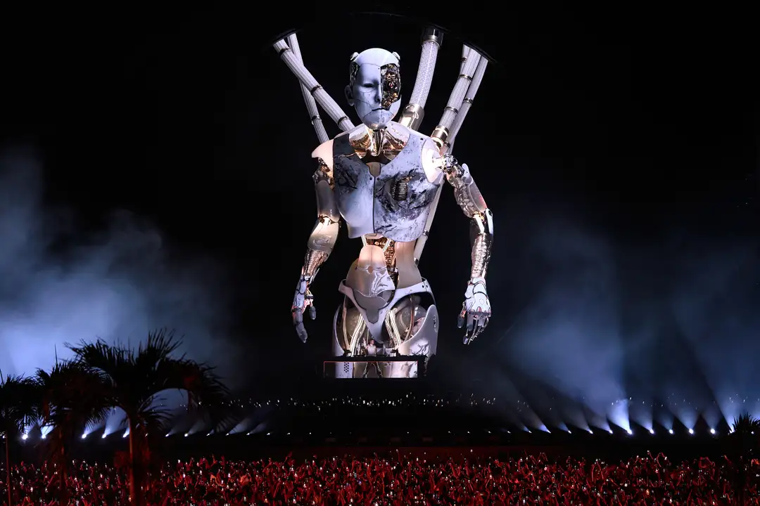 Giant holographic figure with raised arms projected above a crowded nighttime venue with palm trees visible on the sides