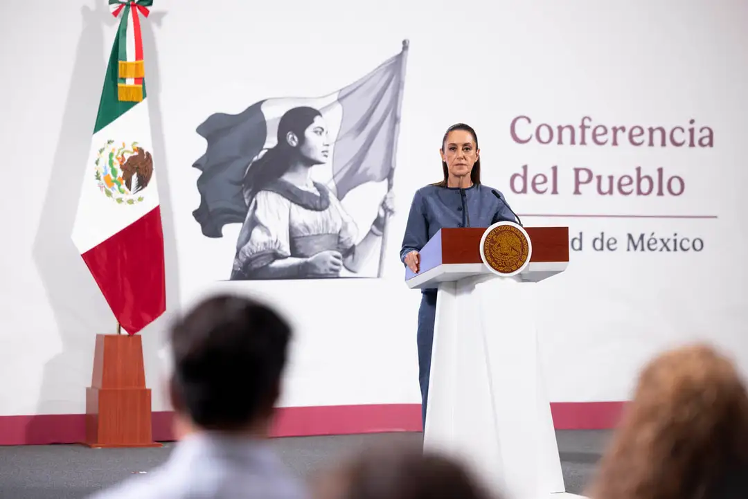 Woman speaking at a podium during a press conference with Mexican flag and mural behind her