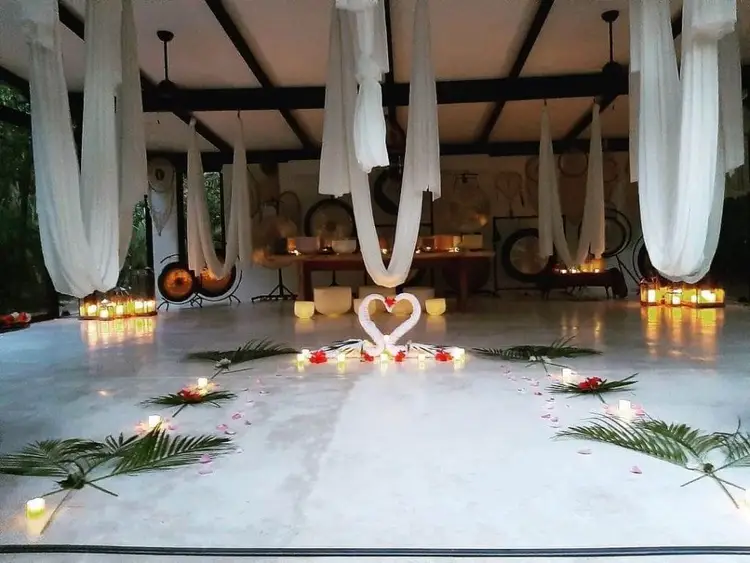 A covered open-air space decorated with white draped fabric, candles, and a heart-shaped floral arrangement on the floor surrounded by palm fronds and petals A covered open-air space decorated with white draped fabric, candles, and a heart-shaped floral arrangement on the floor surrounded by palm fronds and petals