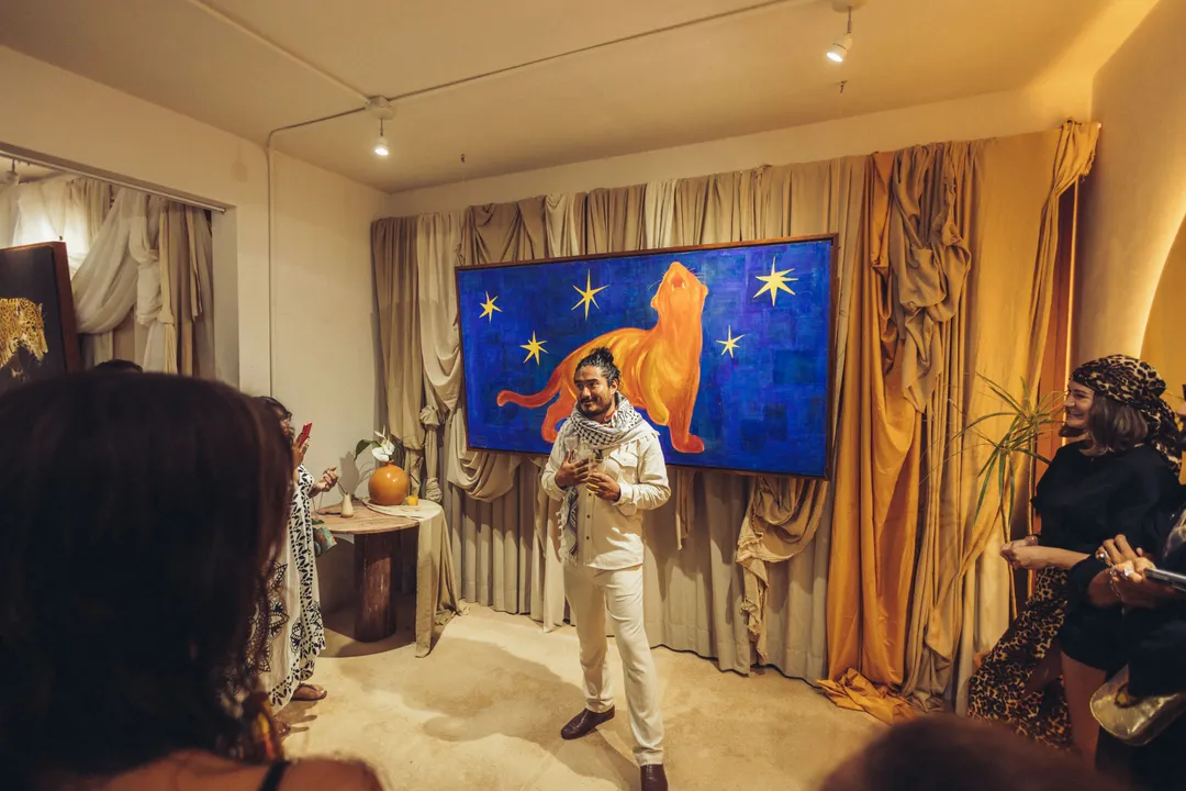 Man in cream-colored suit speaks to gathered guests in an interior space with gold curtains and a blue artwork featuring a lion and stars on the wall