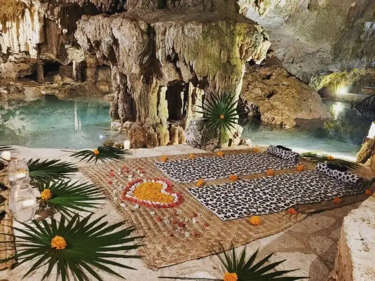 Decorated mats lie on a cave floor beside turquoise pools, surrounded by stalactites and tropical plants with yellow flowers Decorated mats lie on a cave floor beside turquoise pools, surrounded by stalactites and tropical plants with yellow flowers