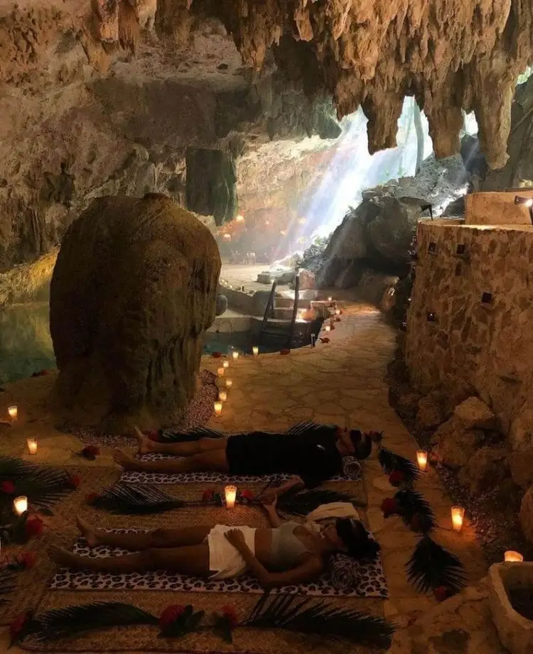 People lie on woven mats surrounded by candles inside a cave with stalactites and a waterfall visible in the background People lie on woven mats surrounded by candles inside a cave with stalactites and a waterfall visible in the background