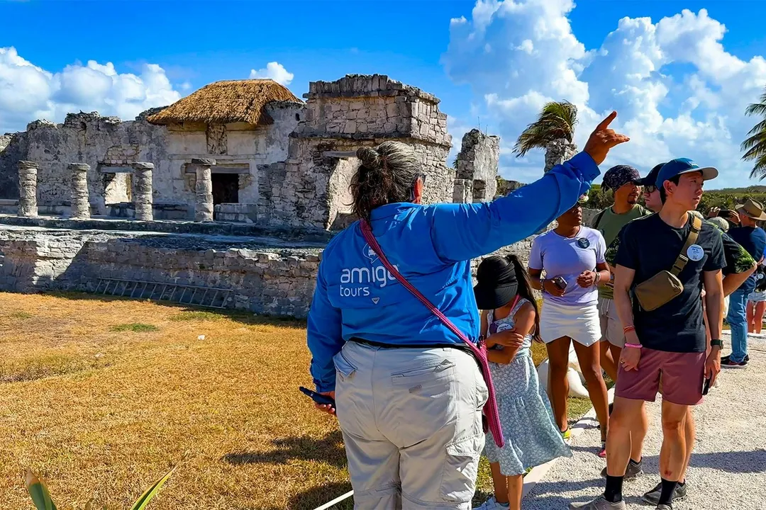 Tour guide in blue jacket gestures while leading a group of visitors at a stone archaeological site with ancient structures and palm trees