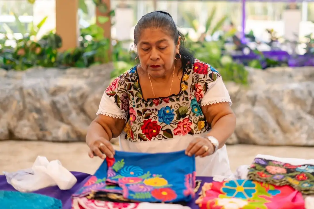 Cheaper Jaguar Park entry may be reaching inland communities Woman in embroidered traditional clothing folds colorful handmade textiles at an outdoor work table surrounded by plants