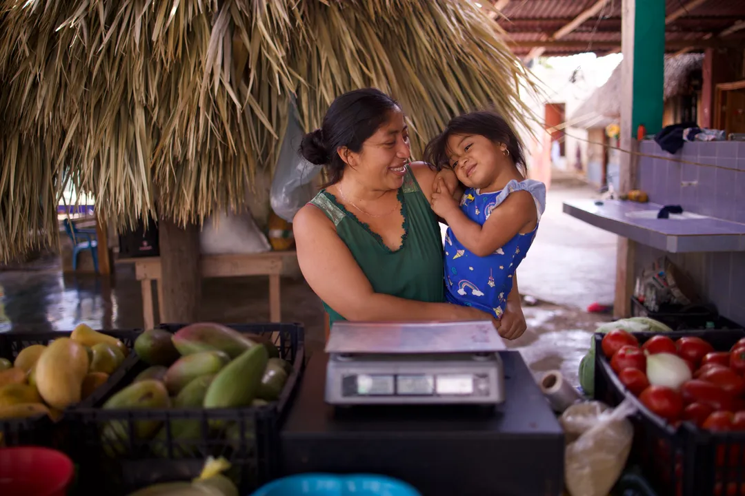 Woman holding a young child at a market stall surrounded by fresh produce and a scale