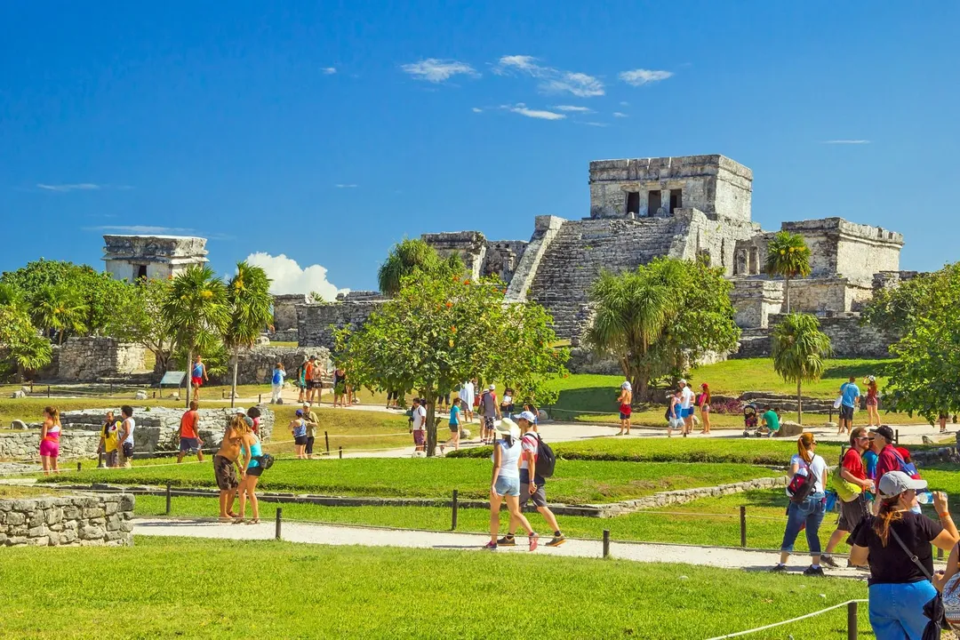 Visitors explore grassy grounds surrounding stone ruins with palm trees and clear blue sky