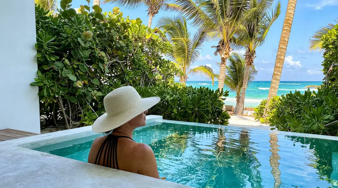 Person wearing a white hat sits in a pool overlooking a tropical beach with palm trees and ocean