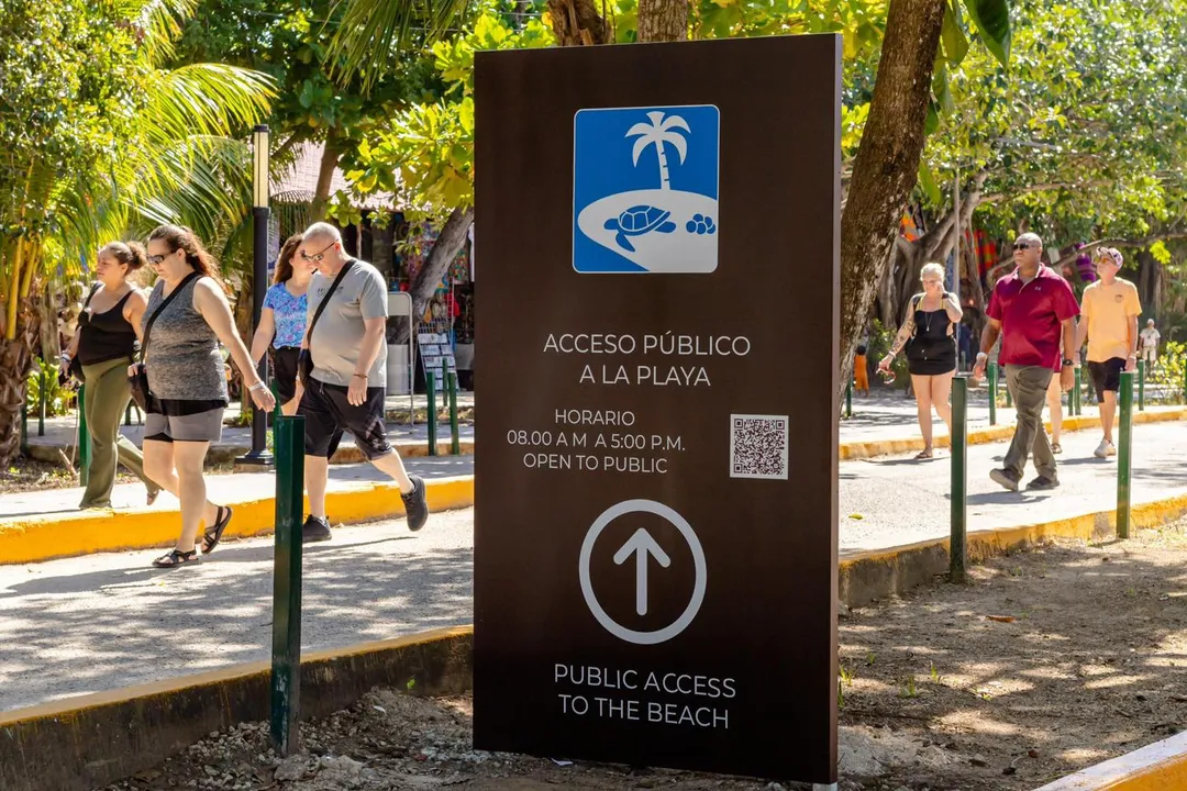 A dark sign with bilingual text and a palm tree logo stands on a tree-lined path where pedestrians walk toward a beach access point