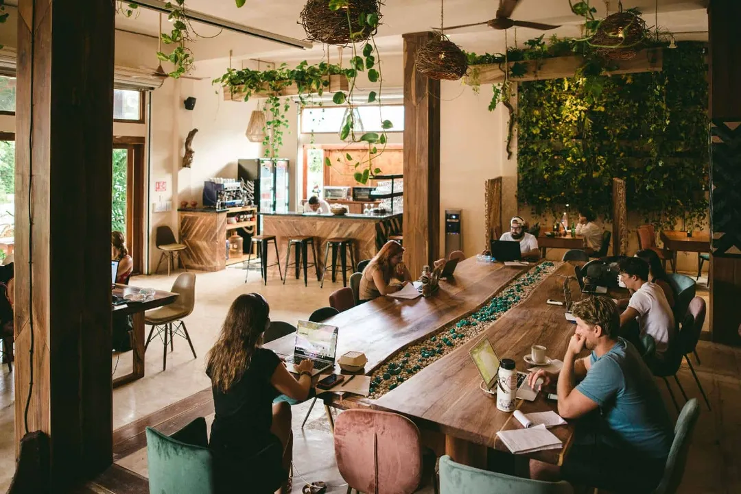 People work at tables and desks in a plant-filled coworking space with wooden furniture and hanging greenery