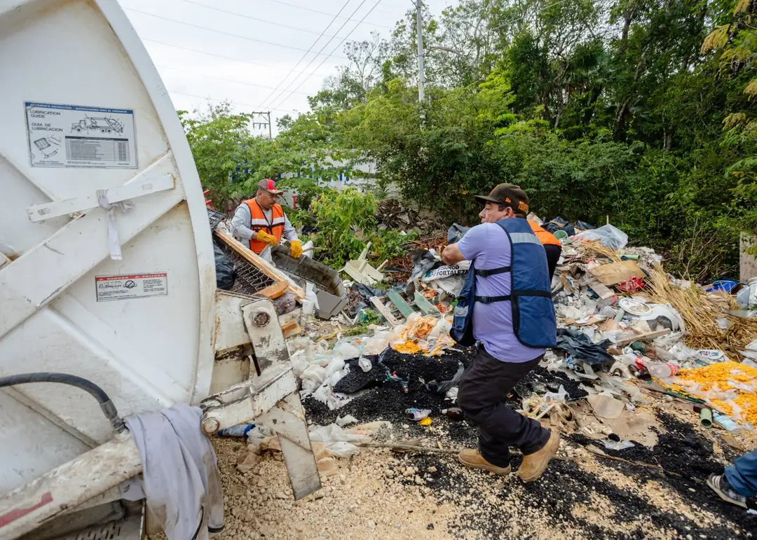Workers in safety vests sort through piles of waste and debris at an illegal dumping site surrounded by vegetation