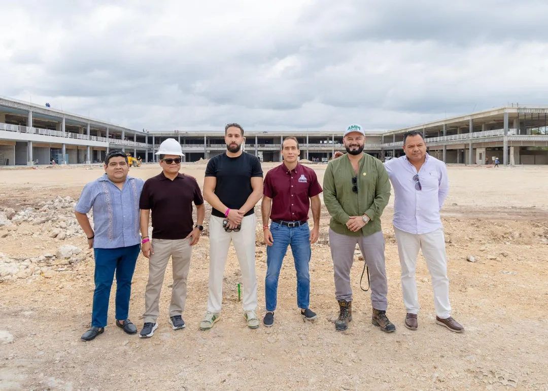 Tulum Gets Its First Commercial Plaza as Centro Tulum Prepares to Open With 60 Stores and 900 Jobs Six men stand in a line on a construction site in front of a large multi-story building under development