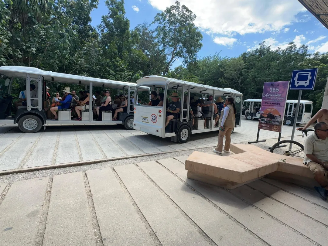 Tulum Tourists Denied Entry at Parque del Jaguar as Rules Clash Tour tram with passengers stopped at an outdoor station near signage, with visitors waiting and wooden seating in the foreground