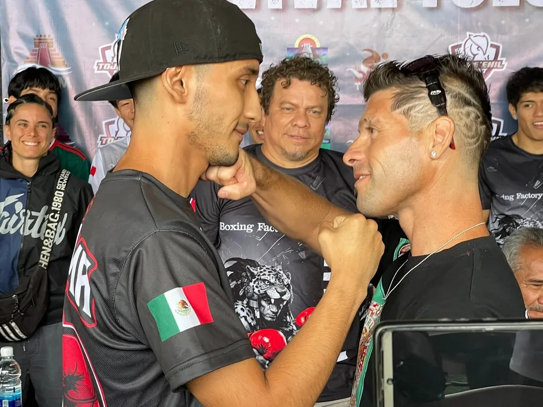 Two men face each other in a confrontational pose at a boxing event, with spectators and a branded backdrop visible behind them