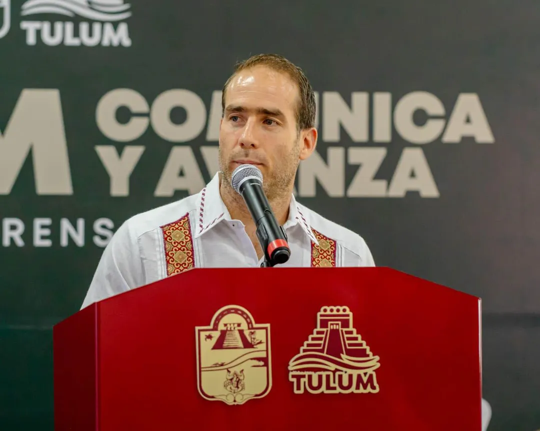 Man wearing white shirt with embroidered vest speaks at red podium with Tulum logo during a presentation or press event