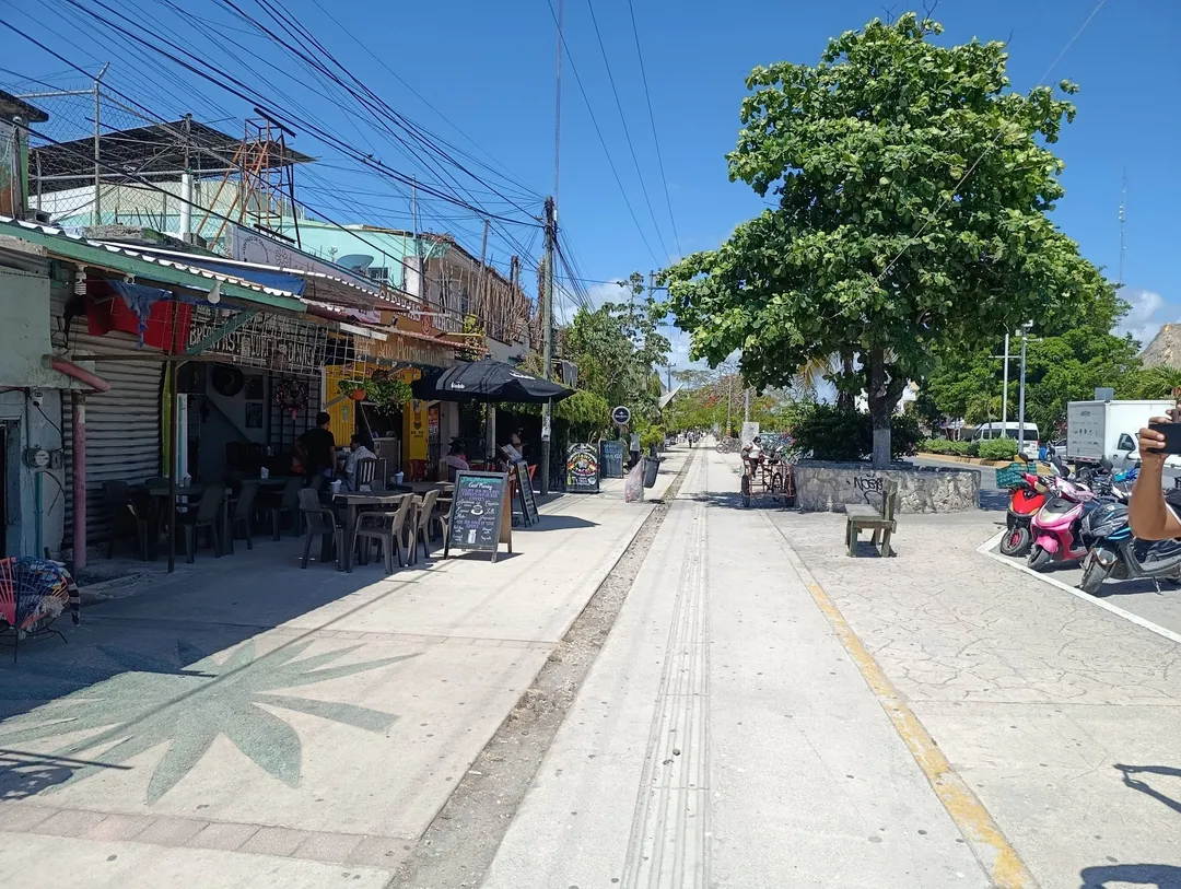 A street lined with small shops and restaurants under power lines, with motorcycles parked on the right side and a large tree in the distance on a clear day