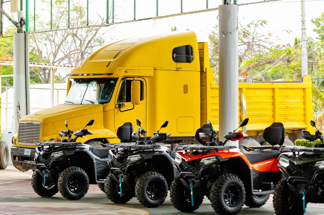 Tulum collects 2,458 tons of sargassum in 2026 as new equipment joins beach crews Yellow semi truck and trailer parked behind a row of black and orange all-terrain vehicles under a covered structure