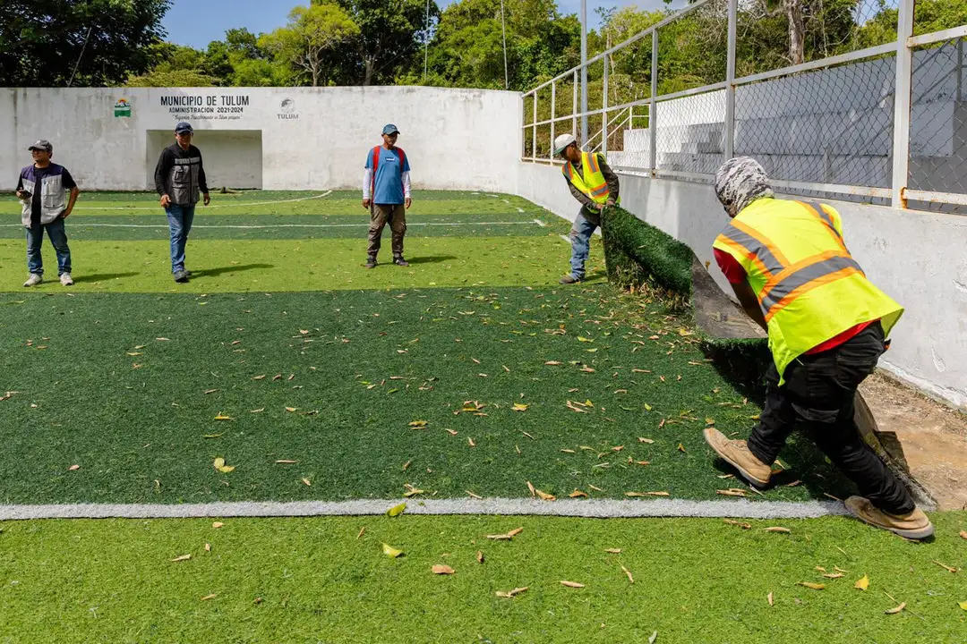 A person in a yellow safety vest demonstrates a move on artificial turf while others watch at a sports facility with white walls and metal fencing
