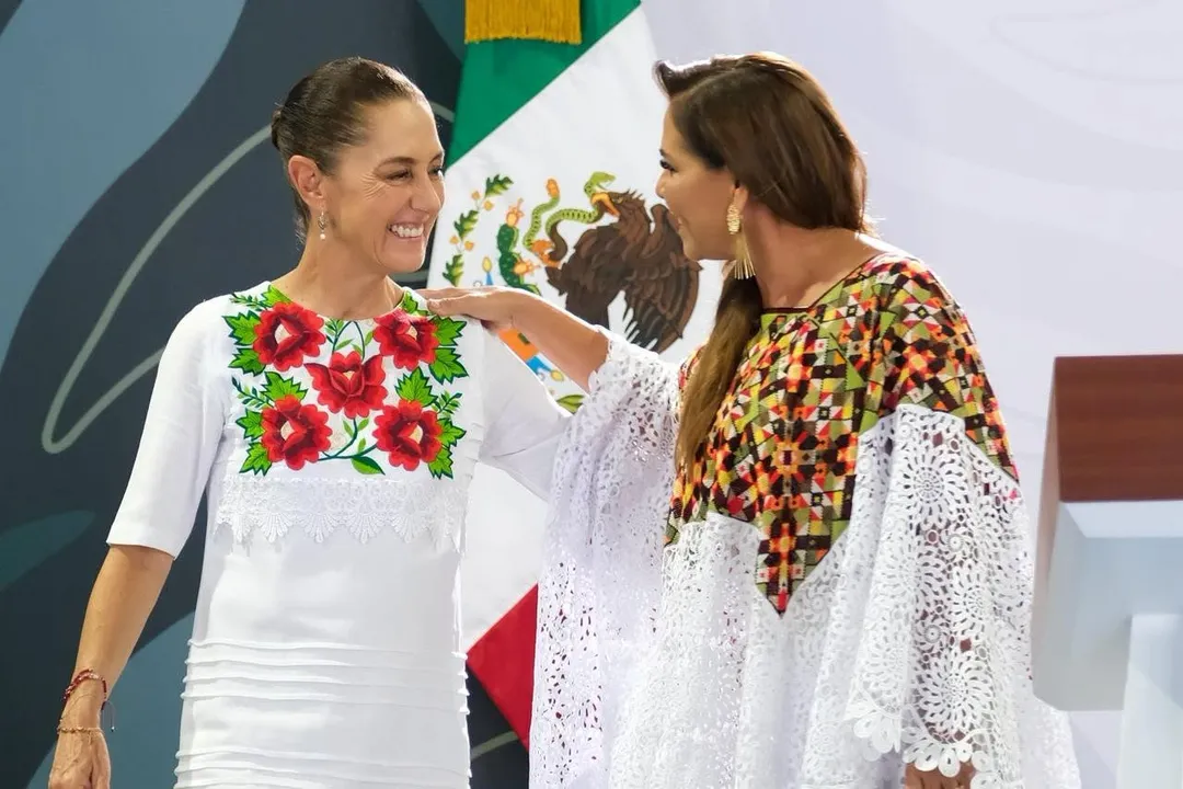 Two women in traditional embroidered white clothing stand together in front of a Mexican flag, smiling at each other during what appears to be an official event